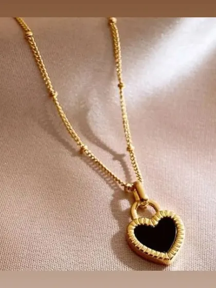 A unique heart-shaped padlock pendant with a black inlay and textured gold-toned border, on a beaded chain. A modern and edgy design.