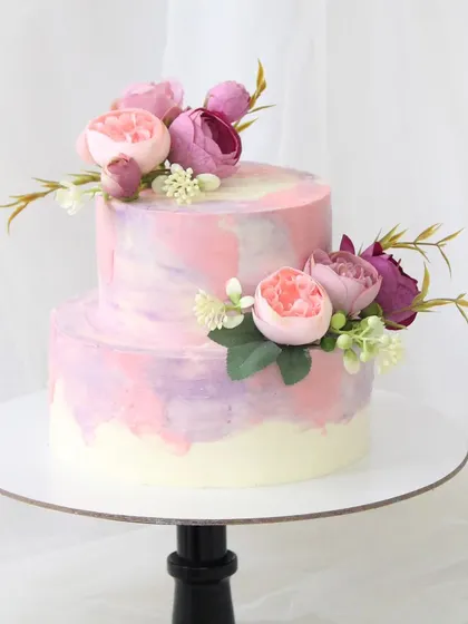A lovely two-tiered cake with a pink, purple, and mauve watercolor effect. The design is beautifully balanced with arrangements of fresh peonies on each tier.