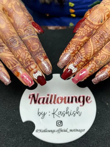 A rich and warm bridal nail set. This design combines deep red and pink nails with glitter accents and rhinestone bows, perfectly matching the intricate mehendi.