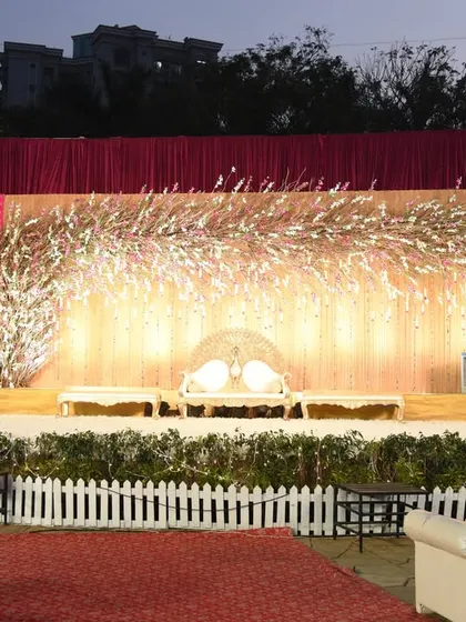 This wedding reception stage features a unique design with golden-white artificial branches against a shimmering backdrop. The white picket fence adds a charming touch to the guest seating area.