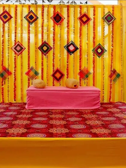 A classic Haldi or Mehndi stage with a backdrop of yellow drapes and marigold strings. The simple seating on a patterned rug is perfect for a traditional home ceremony.