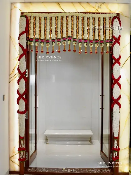 A pristine white marble pooja room entrance adorned with traditional red and white floral garlands. The contrast creates a very elegant and divine look.