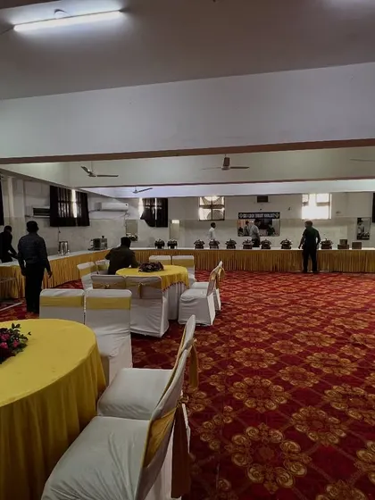 A wide view of a banquet hall during setup, showing the arrangement of multiple tables, chair covers, and the buffet station in the background.