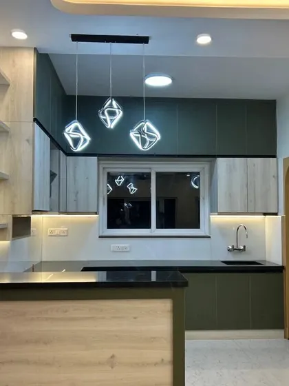 An open kitchen with a breakfast counter that separates it from the living area. The design uses a mix of olive green and light wood-finish cabinets, complemented by modern geometric pendant lights.