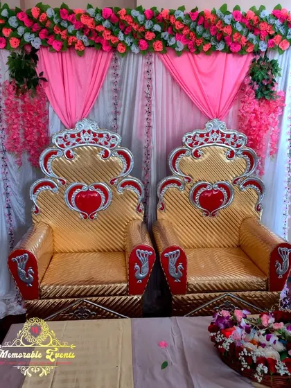 A simple yet elegant engagement decoration for a home setting, featuring ornate golden chairs against a pink and white draped backdrop with floral accents.