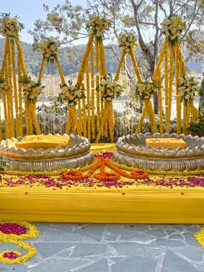 A traditional Haldi seating arrangement with two lotus-shaped brass urlis on a yellow stage, decorated with marigold garlands.