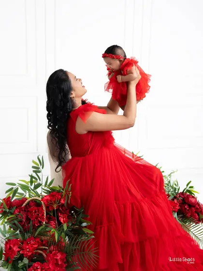 A vibrant and joyful portrait of a mother and her newborn daughter. Dressed in stunning matching red gowns, the mother lifts her baby up in celebration, surrounded by beautiful red flowers.
