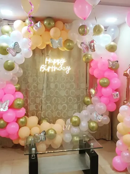 A beautiful first birthday decoration with a circular balloon arch in pink, peach, and gold. The design is accented with silver butterfly cutouts and a 'Happy Birthday' neon sign for a whimsical touch.