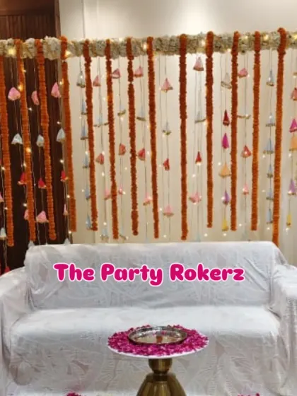 A traditional backdrop for a home kirtan. The decor consists of a seating area with a backdrop of marigold and bell strings, illuminated with fairy lights.