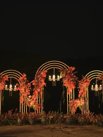 The same welcome dinner setup illuminated at night. The lighting transforms the space, making the red florals and golden arches glow for a dramatic and romantic effect.