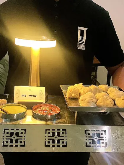 A server presenting a tray of assorted snacks, including veg momos with dipping sauces.