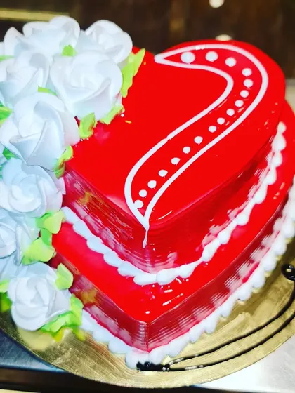 A beautiful heart-shaped cake with a vibrant red glaze, perfect for anniversaries or Valentine's Day. It's elegantly decorated with a cascade of white frosting roses.