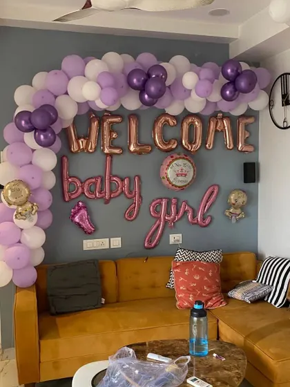 A 'Welcome Baby Girl' decoration in a living room. The setup includes a purple and white balloon arch and rose gold foil letters.