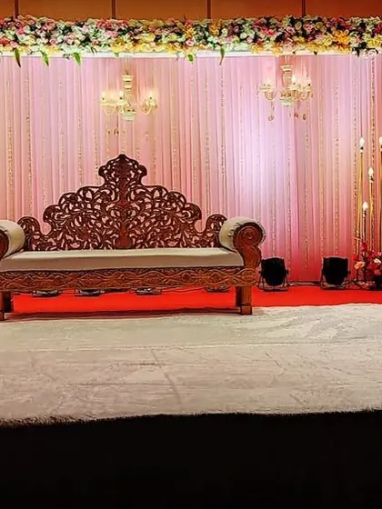 A classic wedding stage design. The carved wooden sofa is set against a backdrop of pink curtains, floral borders, and candle stands, creating a warm and traditional look.