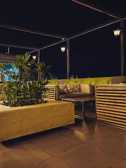 A cozy lounge corner on the rooftop, featuring slatted wood sofas and a large planter. The overhead pergola with hanging lanterns provides structure and ambient lighting.