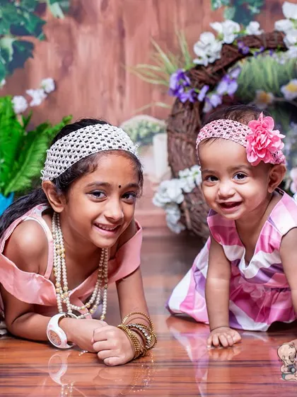 Two sisters lie on the floor in a floral studio setting, smiling for the camera. Their relaxed and happy mood makes this a charming and lovely sibling photograph.