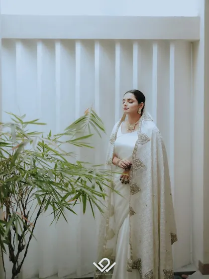 A serene portrait of a Muslim bride in a white ensemble, standing near a bamboo plant against a minimalist white wall.