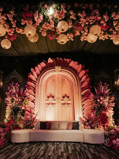 A stunning stage or photo backdrop featuring a large, pink arched doorway framed by feathery petals. The area is flanked by massive floral arrangements and topped with a ceiling of hanging flowers and lanterns.