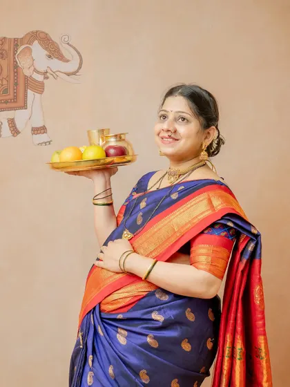 A portrait of the mother-to-be looking up with a hopeful expression, holding a thali of fruits. The elephant motif in the background adds a touch of traditional artistry to the scene.