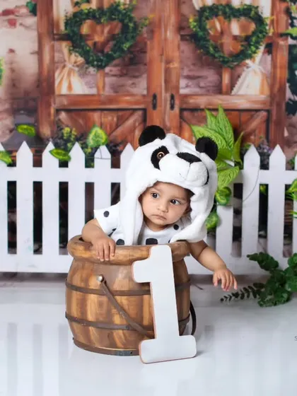 A cute baby wearing a panda hat and peeking out of a wooden barrel for her first birthday photos.