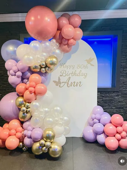 An elegant 80th birthday backdrop for Ann. The setup features an arched panel and a beautiful balloon arrangement in pastel pink, purple, and white with gold accents.