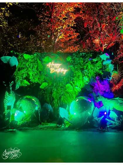A wider shot of the neon photo wall, showing how the installation is set against the natural backdrop of the trees, blending the artificial and natural elements.