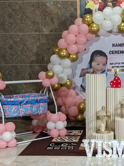 A naming ceremony setup featuring a decorated cradle, a pink and gold balloon arch, and a personalized backdrop.