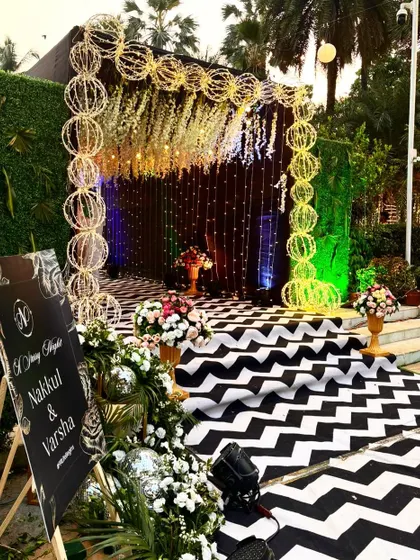 A unique photo booth or entrance for a modern reception. The design uses a black and white chevron carpet, a curtain of string lights, and an arch made of illuminated spheres.