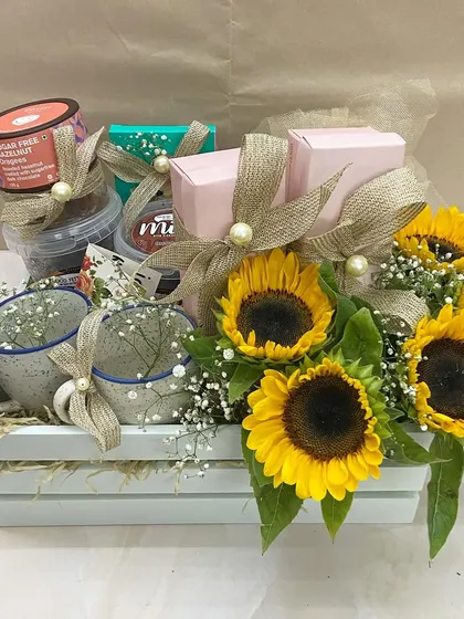 A rustic and sunny surprise basket for Mother's Day. This white wooden crate is filled with coffee mugs, treats, and a cheerful bouquet of fresh sunflowers.