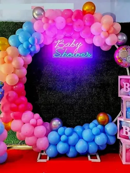 A vibrant and colorful baby shower setup against a green hedge wall. The balloon ring uses a rainbow of colors, and the "Baby Shower" neon sign glows brightly, making it a great spot for photos.