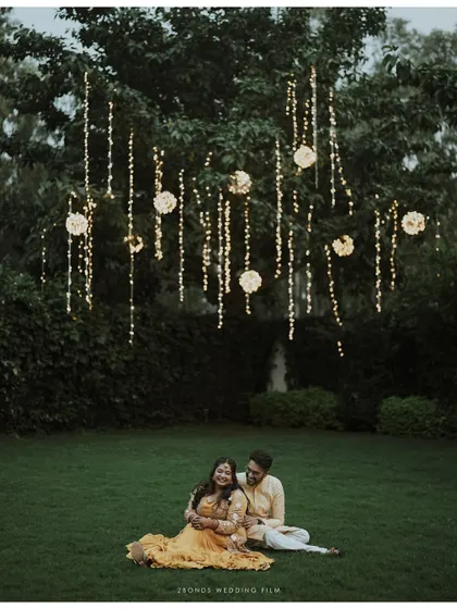 A magical evening portrait of the couple sitting on the grass under a tree adorned with beautiful hanging lights.