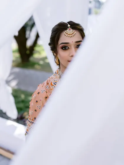 A creative and artistic shot of the bride, peeking through white curtains. The focus is on her expressive eyes, which are beautifully defined to complement her traditional attire.