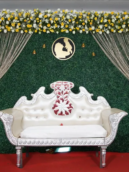 A clean and modern baby shower stage with a green hedge backdrop, accented with a string curtain and a floral border of yellow and white flowers.