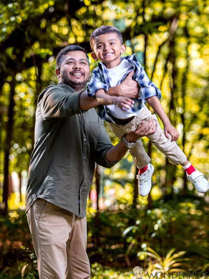 A father joyfully lifts his son in the air while walking through a park. The genuine happiness on both their faces makes this a perfect candid shot of fatherhood.
