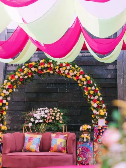 A colorful Mehendi stage with a pink sofa set against a floral arch and a backdrop of pink and lime green drapes.