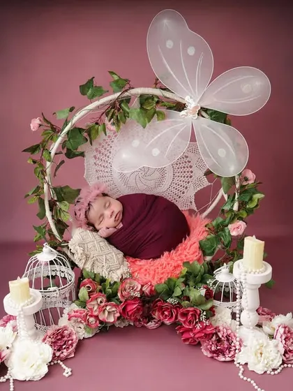 A magical, fairytale scene. This wide shot shows the newborn sleeping in a floral hoop with butterfly wings, surrounded by candles and birdcages for an enchanted forest feel.