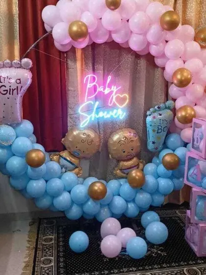 A 'He or She' themed baby shower with a circular balloon arch in pink and blue. The decor includes a neon sign and foil balloons for 'It's a Boy' and 'It's a Girl'.