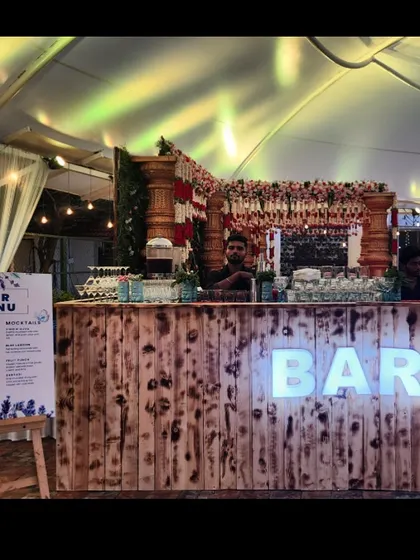 A rustic bar counter set up for a wedding event under a tent. The bar is decorated with traditional floral garlands, and a large, custom-printed bar menu is displayed on an easel for guests to see.