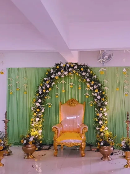 A serene and elegant Valaikappu or Seemantham setup. The decor features a graceful arch of fresh yellow and white flowers, soft green drapes, and traditional brass lamps for a blessed occasion.