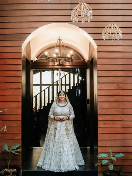 A grand entrance shot. The bride stands framed in a large wooden doorway, her stunning white lehenga filling the frame, creating a moment of regal elegance.