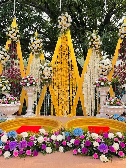 The main seating for the Haldi ceremony. Two large brass urlis are filled with flowers, set against a backdrop of geometric panels and cascading marigold strings.