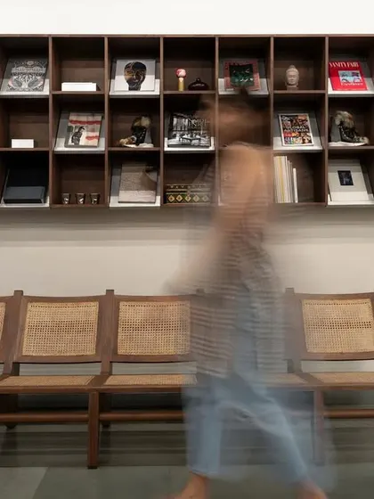 A detail of the waiting area in the industrial-chic fashion showroom. A traditional wooden bench with cane weaving provides a warm, organic contrast to the modern, minimalist library of design books and objects above it.