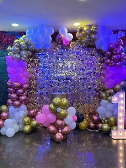 A glamorous first birthday setup with a gold sequin wall, a "1" marquee number, and a colorful balloon arch in pink, purple, and gold.