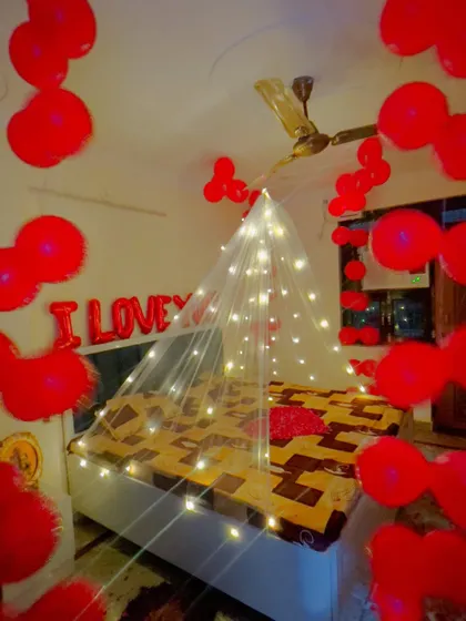 A wider view of the red-themed romantic room decor, showing the balloons on the ceiling and the heart-shaped petal arrangement on the bed.