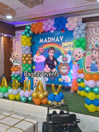A wide view of the Cocomelon themed stage decoration for Madhav's first birthday party. The setup includes a large backdrop, character cutouts, and balloon letters spelling out his name.