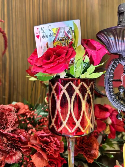 A close-up of a red patterned glass holding a small bouquet and playing cards, a fun and quirky detail for a themed event.