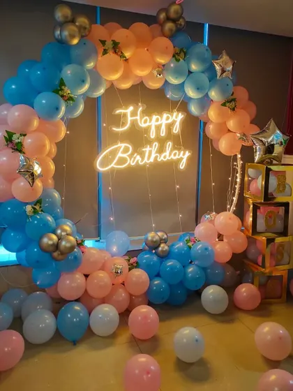 A classic circular balloon arch in a playful pink, blue, and peach color scheme. The "Happy Birthday" neon sign in the center adds a warm glow, making it a great backdrop for cake cutting and photos.
