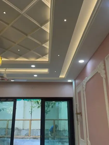 A close-up of an intricate false ceiling with a coffered design and a large, elegant light fixture. The pink walls with decorative molding add a touch of classic European style.