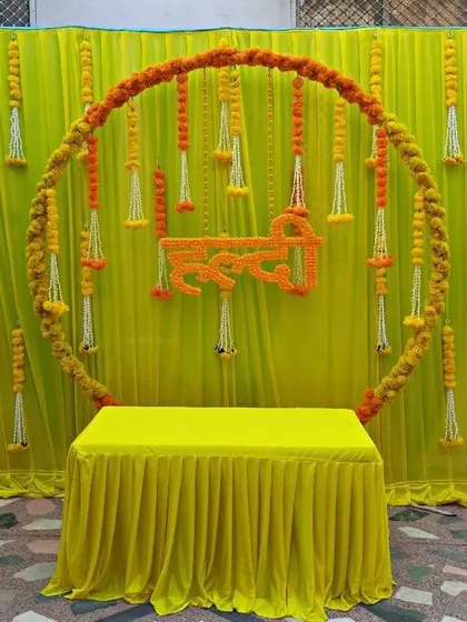 This simple yet elegant Haldi ceremony setup features a circular frame wrapped in marigold garlands against a yellow curtain. The hanging floral tassels add detail to this clean and traditional design.