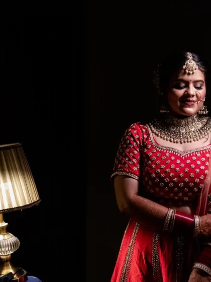 A beautifully lit bridal portrait. The bride stands beside a vintage lamp, creating a warm and intimate atmosphere that contrasts with the dark background, making her red lehenga pop.